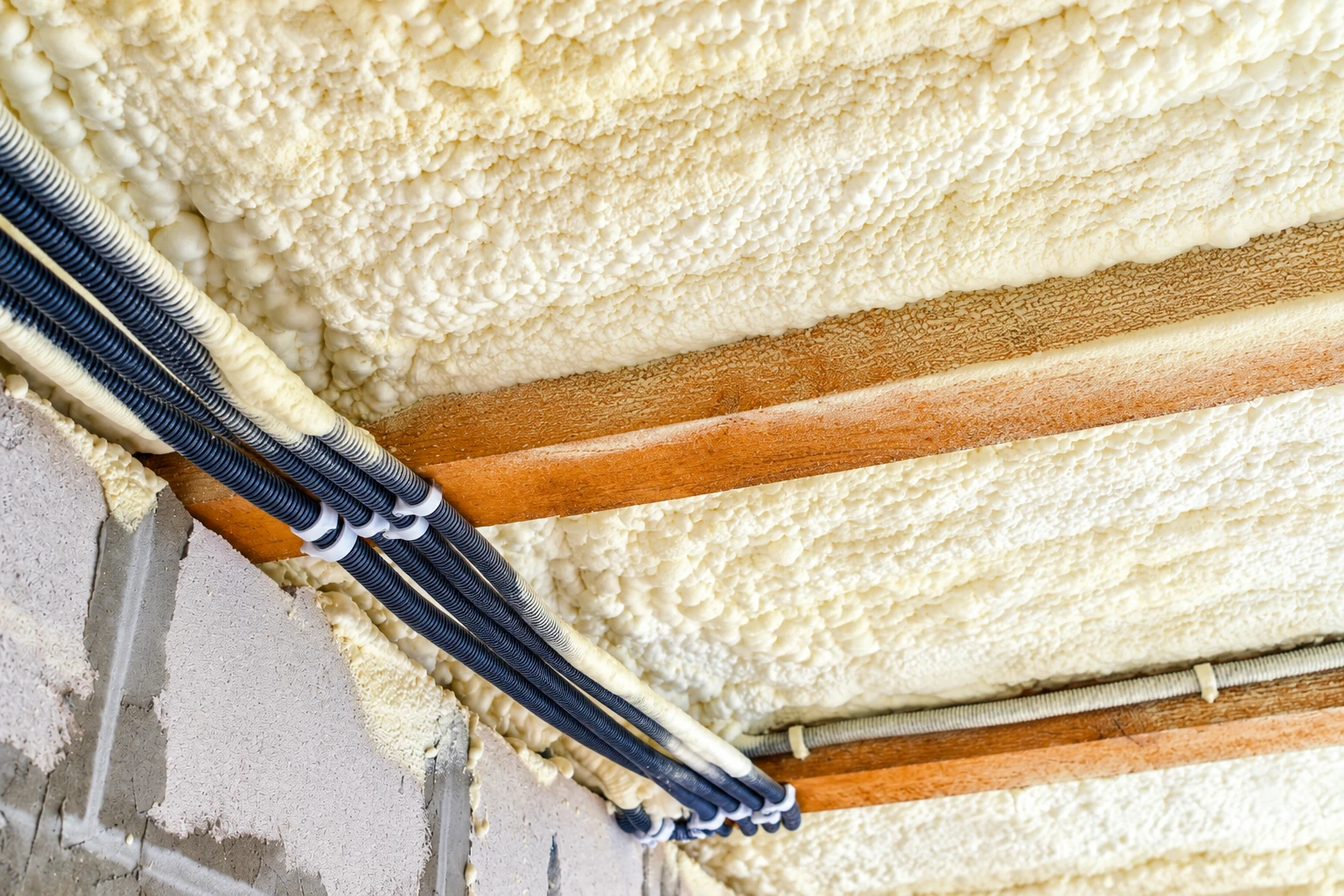 Thick white spray foam coats an attic ceiling between wooden beams, beside bundled cables and a block wall.
