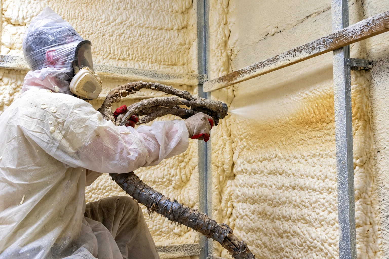 A worker in protective coveralls and a respirator sprays foam insulation onto an interior wall framed with metal studs.