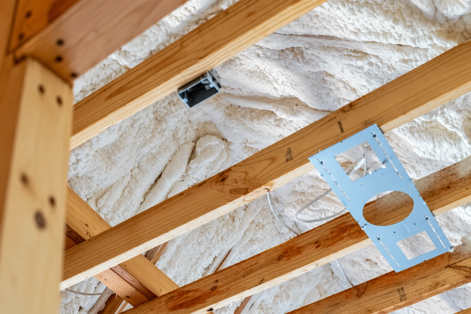 White spray foam fills the spaces between exposed wooden roof rafters, with a blue mounting plate and wiring visible.